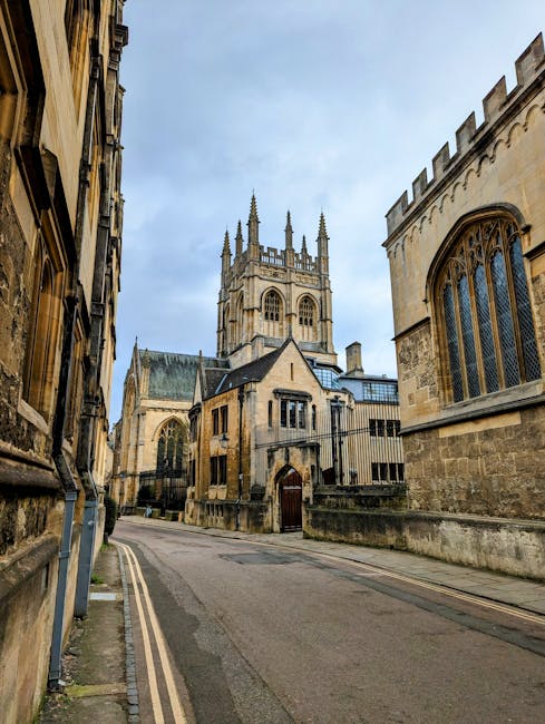 This image shows a narrow street in a historic town with old stone buildings on either side, leading towards a large medieval-style church with tall spires and intricate gothic windows. The street is paved with asphalt and has a double yellow line along the curb. There are no vehicles or pedestrians visible. The scene is captured during daylight under a partly cloudy sky, with diffused natural light illuminating the stone facades. The setting appears quiet and typical of a European town centre, with the church being a prominent landmark. Man with Van Merton, a professional removals service, might utilize such locations for efficient house relocation or furniture transport, ensuring careful handling of bulky items close to historic buildings and narrow urban streets.