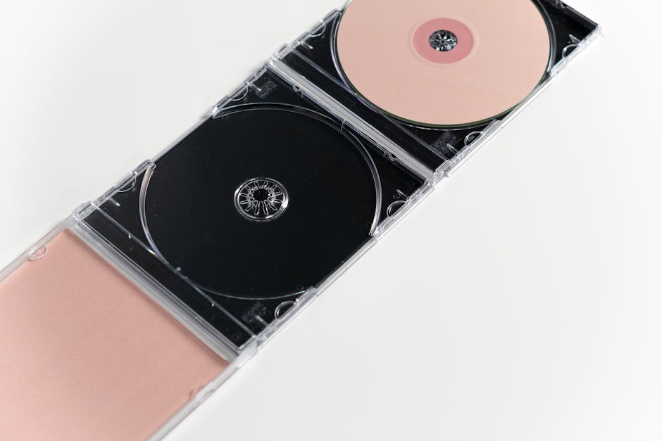 A close-up photograph of a transparent plastic CD case containing two compact discs; one CD is black and the other is a light pink color. The case is placed on a plain white surface, and the discs are securely seated within the cases, showing their central hubs and reflective surfaces. In the background, partially visible, are packing materials such as foam padding or cushioning, which are used for protecting media during home relocation or furniture transport. The environment appears to be indoors with soft, even lighting, highlighting the clarity of the plastic case and the details of the discs. This image relates to packing and moving processes, as well as media or electronic items consolidation, relevant to professional removals services like those offered by Man with Van Merton, especially for house moves involving careful packing of media collections.