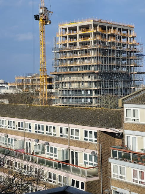 A multi-storey residential building under construction, surrounded by scaffolding and yellow safety barriers, with a large yellow tower crane positioned to the left of the building. In the foreground, there are traditional low-rise apartment blocks with white-framed windows and terraced roofs. The construction site is set against a clear blue sky, and the scene depicts the ongoing development process typical of urban house and flat relocations, as managed by companies like Man with Van Merton. This image highlights the scale of project logistics involved in moving or renovating properties, showcasing construction equipment and building materials in a residential environment.