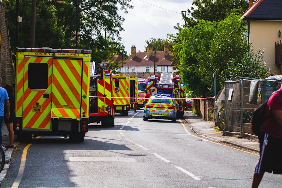 A street scene in Merton during an emergency response with multiple emergency vehicles, including a London ambulance and police cars, parked along the road. The ambulance, marked with high-visibility yellow and red chevrons, is positioned near the sidewalk, while police vehicles with flashing blue lights are nearby. Several fire trucks and personnel, dressed in protective gear, are present in the background, potentially engaged in coordinated efforts. The street is sectioned off with red and white tape, indicating an active response area, and a few bystanders are observing from the sidewalk. Trees line the street, with residential houses visible in the distance, featuring pitched roofs and chimneys. The scene highlights the logistical complexity of emergency situations that can impact local urban areas, often involving coordinated transport and containment efforts by emergency services, akin to those in house removals or relocation projects undertaken by companies like Man with Van Merton.