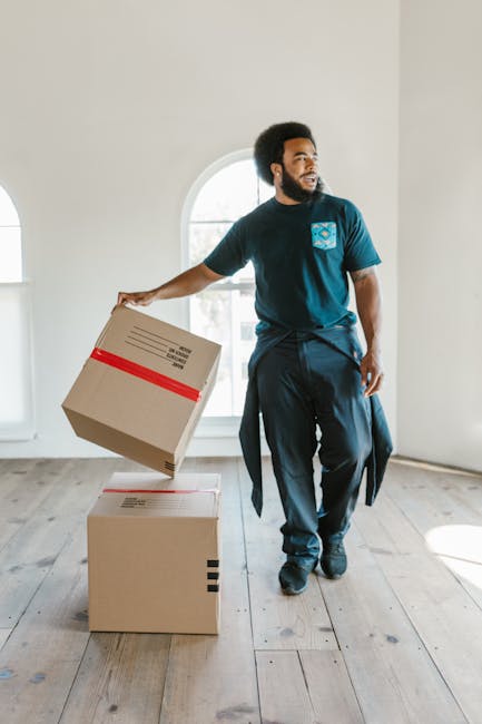 A man with dark curly hair and a beard, wearing a blue t-shirt with a printed logo and dark trousers, is standing inside a bright room with a large arched window in the background. He is holding a cardboard box wrapped with red tape in one hand, preparing to place it on top of another cardboard box positioned on a light wooden floor. Several similar boxes are stacked next to him, indicating a moving or packing process related to house relocation, with the equipment and environment suggesting professional furniture transport and packing activities. The natural light from the window illuminates the scene, emphasizing the organized and efficient setup for a home removal or moving service, as performed by [COMPANY_NAME].