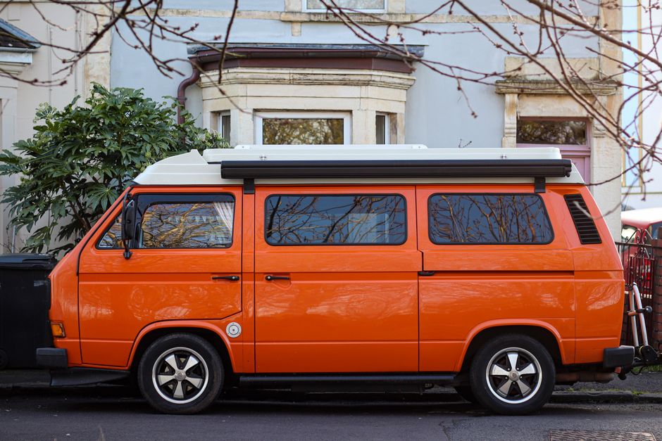 A side view of an orange Volkswagen camper van parked on the street in front of a residential property with a bay window and a doorway. The van is equipped with a white roof rack, and the windows reflect nearby leafless trees. The vehicle is positioned close to the curb, with the driver’s side door visible, and appears to be involved in a home relocation or furniture transport process, as part of professional removals services by Man with Van Merton. There are cardboard boxes and other packing materials visible inside the van through the windows, indicating packing and moving activities. The setting suggests a clear day with natural daylight illuminating the scene, and the environment includes a bicycle secured to the back of the van, emphasizing the practical aspects of moving logistics, including parking permits and urban transport considerations within Merton.
