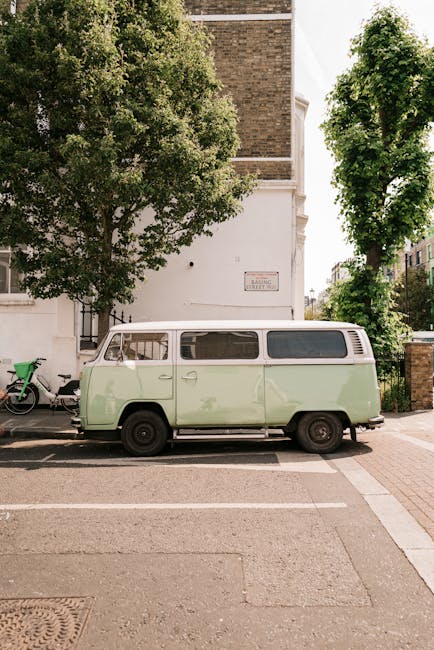 A vintage light green and white Volkswagen Type 2 van is parked on the side of a quiet street adjacent to a white building with a brick upper section, flanked by two large leafy trees providing partial shade. The van's side windows are visible, along with its black tires and simple wheel hubs. In the background, a green bicycle is secured to a metal rack near the building, which is partially obscured by foliage. The street surface is paved, with white parking lines indicating designated parking spaces. The scene captures a clear, sunny day with natural light illuminating the vehicle and surroundings, aligning with context for house removals and relocating services offered by Man with Van Merton. The setting suggests an area suitable for home relocation activities, including furniture transport and packing preparations, within urban residential surroundings.