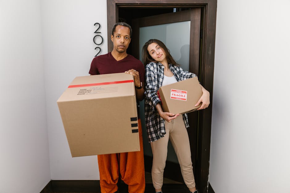 A man and a woman stand in a narrow hallway outside a door, each holding cardboard moving boxes; the man holds a large box with black tape and labels, while the woman holds a smaller box marked 'Fragi