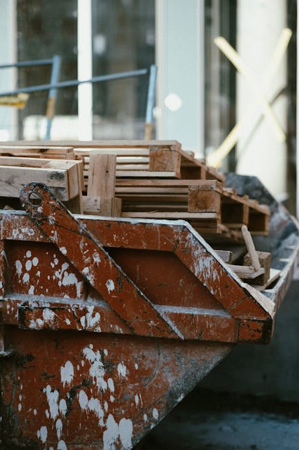A close-up view of a rusty, weathered boat positioned on the pavement outside a building with large glass windows, partially filled with wooden pallets and other packing materials. The pallets are sta