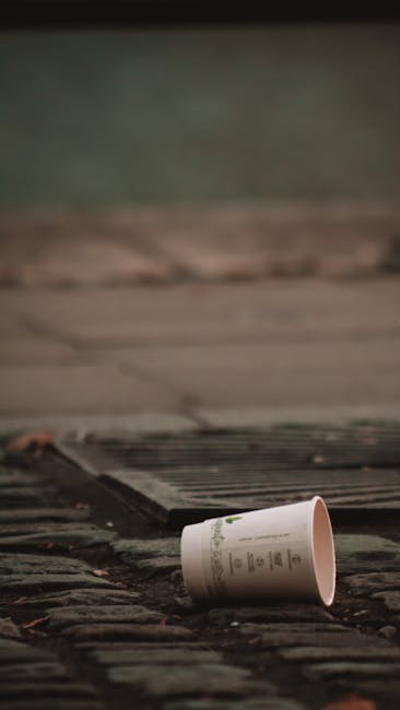 A close-up view of a rusty, weathered boat positioned on the pavement outside a building with large glass windows, partially filled with wooden pallets and other packing materials. The pallets are sta
