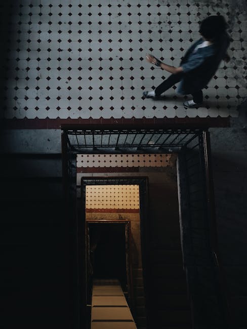 A top-down view of a person in a dark corridor of a multi-storey building, carrying a mobile device while ascending a staircase with metal railings. The staircase has a tiled wall with a small, circular pattern in red and white, visible from the landing to the lower floor. Part of the person's outfit includes dark trousers and a blue shirt or jacket. The environment appears to be in a residential or commercial property, possibly during a house or apartment move, with minimal lighting emphasizing the staircase’s depth and the person's upward movement. This scene illustrates a typical stair-only home relocation process, with no furniture or boxes visible but a focus on moving within the property. Man with Van Merton provides removals services for such internal moving tasks, including safe stair navigation and efficient packing and transport of belongings.