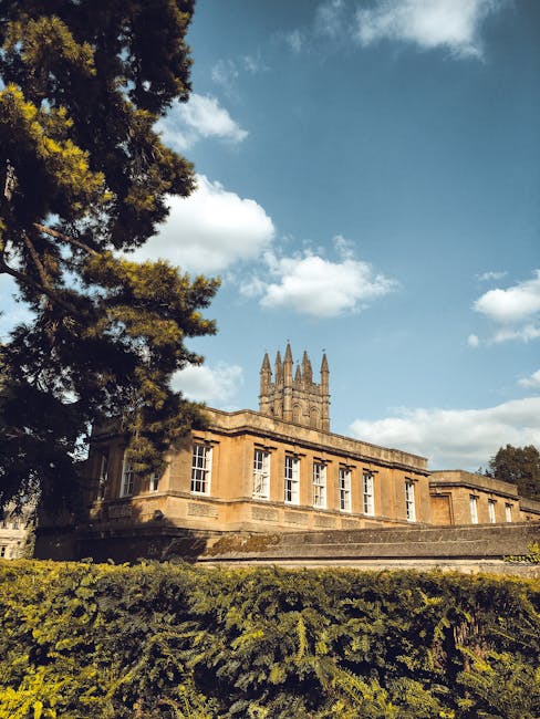 Photograph of a historic stone building with tall windows and a prominent tower with multiple spires, set against a bright sky with scattered clouds. In the foreground, a large evergreen tree with dark green foliage partially frames the left side, while well-maintained hedges run along the base of the building. The scene appears to be taken outdoors during daytime in a scenic area, possibly part of a university or cultural site near Wimbledon Common, with the building’s classic architectural features clearly visible. The setting suggests a peaceful environment suitable for house removals or relocation services, and the image conveys a sense of stability and tradition, aligning with professional moving and packing processes, such as those managed by Man with Van Merton.