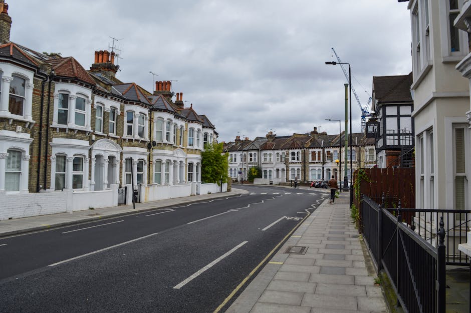 A daytime street scene showing a quiet residential area with Victorian-style terraced houses featuring bay windows and chimneys. The pavement runs alongside the row of homes, with some houses having small front gardens enclosed by black metal fences. Pedestrians are seen walking on the sidewalk, and a crane is visible in the distant background, indicating ongoing construction work. The sky is overcast with grey clouds. This setting reflects a typical London suburb environment suitable for house removals and furniture transport. Occasionally, Man with Van Merton's removal services may be involved in such relocations, providing packing and moving assistance for residents in Wimbledon Hill SW19 and surrounding areas.