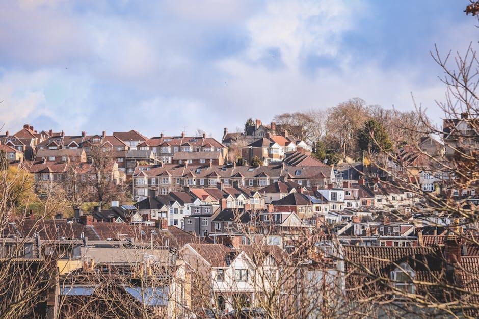 A panoramic view of a residential neighbourhood with numerous terraced and semi-detached houses situated on a hillside under a partly cloudy sky. The foreground features bare tree branches, indicating a season of late autumn or winter. The houses have various roofing styles, primarily tiled in shades of brown and grey, with some dormer windows visible. Behind the houses, a cluster of trees and a clear sky can be seen. This scene exemplifies a typical UK suburban area suitable for home relocations and local removals services. The image, associated with the services of Man with Van Merton, highlights the dense housing layout often involved in furniture transport and packing and moving processes, emphasizing the importance of careful planning in house removals within areas like Wimbledon Hill SW19.